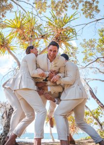 Bridesmaids and groomsmen laughing and posing with Zoe and Deon on the grass by the ocean at their Noosa wedding.