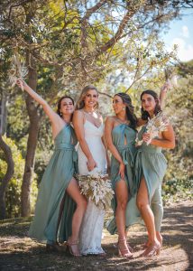 Bridesmaids and groomsmen laughing and posing with Zoe and Deon on the grass by the ocean at their Noosa wedding.