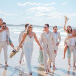 Bridesmaids and groomsmen laughing and posing with Zoe and Deon on the grass by the ocean at their Noosa wedding.