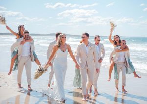 Bridesmaids and groomsmen laughing and posing with Zoe and Deon on the grass by the ocean at their Noosa wedding.