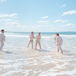 Bridesmaids and groomsmen laughing and posing with Zoe and Deon on the grass by the ocean at their Noosa wedding.