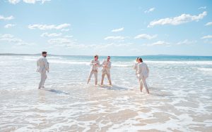 Bridesmaids and groomsmen laughing and posing with Zoe and Deon on the grass by the ocean at their Noosa wedding.