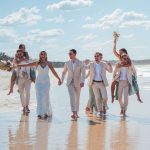 Bridesmaids and groomsmen laughing and posing with Zoe and Deon on the grass by the ocean at their Noosa wedding.