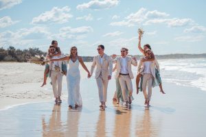 Bridesmaids and groomsmen laughing and posing with Zoe and Deon on the grass by the ocean at their Noosa wedding.