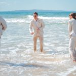 Bridesmaids and groomsmen laughing and posing with Zoe and Deon on the grass by the ocean at their Noosa wedding.